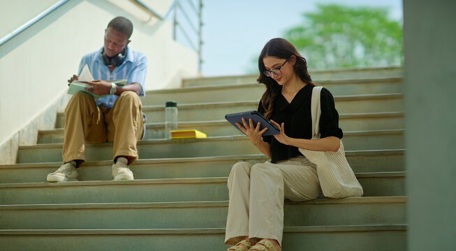 African young adult gen z busy male man sitting on stair hold casual book read self exam notes at place. Indian female girl use tab look screen do work check email learn new day study spend break time