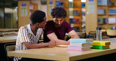 Indian two male pupil sitting on desk table do study talk new project task work share plan ideas young adult gen z boys man guys pair hold pencil look book read exam notes task indoor library place