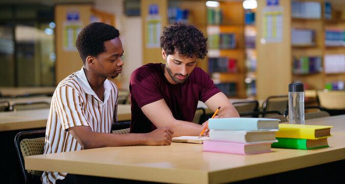 Indian two male pupil sitting on desk table do study talk new project task work share plan ideas young adult gen z boys man guys pair hold pencil look book read exam notes task indoor library place
