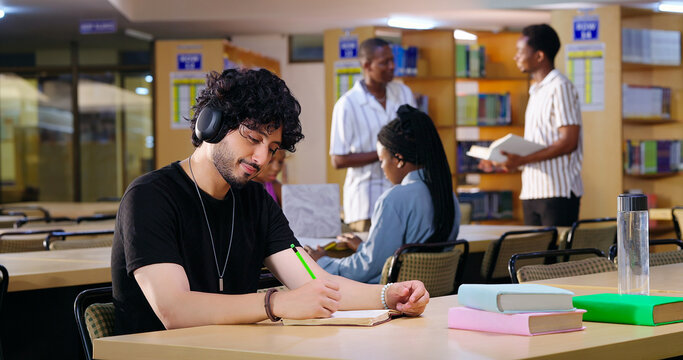 Indian Asian young adult gen z male man calm peace mind student sitting desk table listening music hold pencil write exam notes smart boy do self study read book prepare topics indoor library busy day