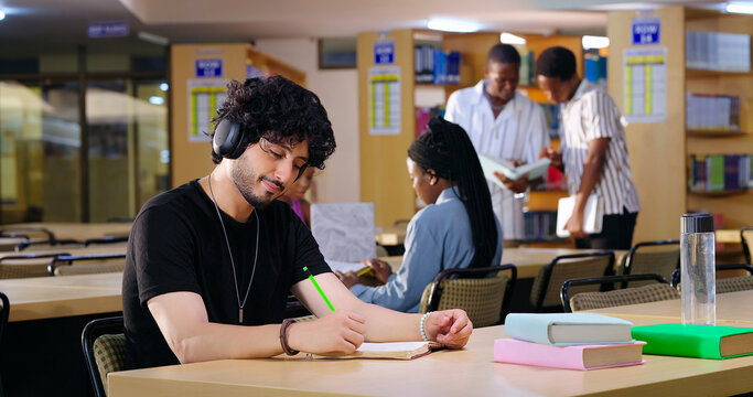 Indian Asian young adult gen z male man calm peace mind sitting desk table listening music hold pencil write exam notes smart boy do self study read book prepare topics indoor library busy day