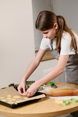 Girl spreads cookie dough on baking sheet, pressing edges, while rolling pin and flour are scattered on table. Teenager enjoys baking cookies, rolling them out, cutting them out with cookie cutters.