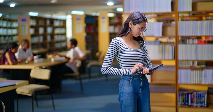 Indian young adult gen z busy woman lady hold tab look screen do online work e learning skill check exam notes. Happy teen girl wear glass candid casual pose enjoy new day time indoor library