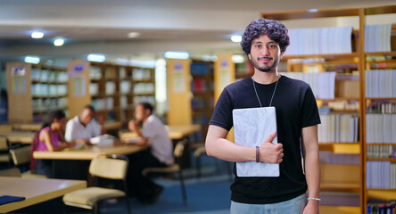 Indian young adult gen z man student standing proud hold new laptop look camera indoor library...