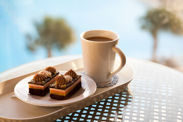 Two cream cakes and a cup of coffee on a tray outdoors, with blurred palm trees and blue water from basin creating a calm, relaxing breakfast or dessert atmosphere.