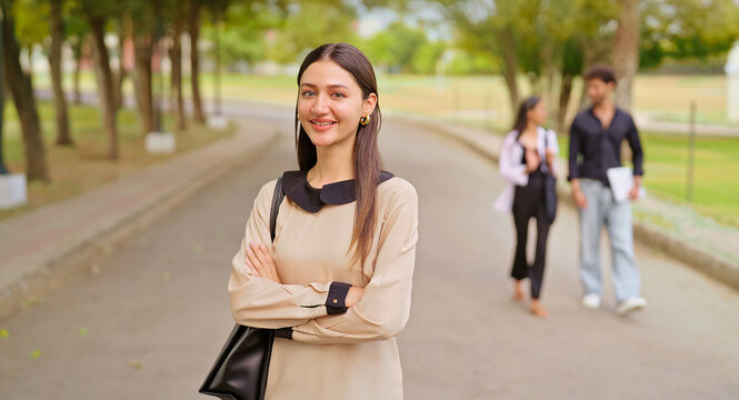 Indian happy young adult gen z proud smart lady female look camera cross arms standing outdoor campus area. Teen age pupil pretty girl fold hand wear bag staring cam do casual pose enjoy new day place