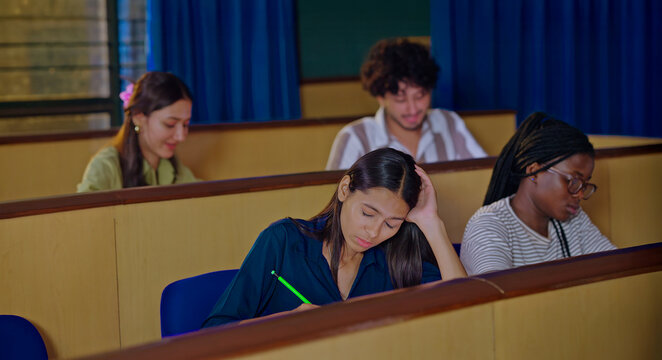 Indian young adult gen z dull pupil girl hold pencil sitting do study writing exam note indoor class room female feel bad tired lazy sleepy have lack energy need rest sleep on desk table college place