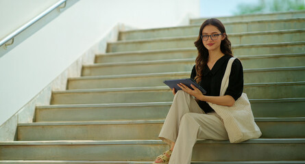 Indian young adult gen z lady woman use tab look screen do study exam check online e work read notes. Teen girl wear glass sitting on stair staring cam causal pose enjoy new day spend break time place