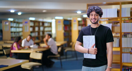 Fototapeta premium Indian asian young adult gen z man confident student standing proud hold new laptop look camera indoor library happy smiling boy guy staring cam do casual pose enjoy joyful day time inside college
