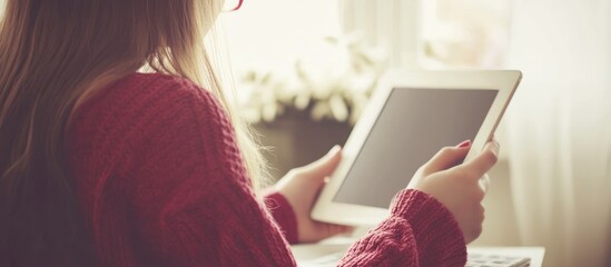 Young woman in a red sweater sits by a window, holding a tablet computer. Sunlight illuminates the scene