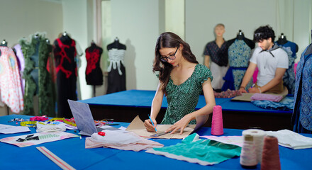 Indian young adult gen z female lady do new day e learning clothe craft course skill indoor class room place teen girl wear glasses use laptop look screen hold pencil fabric make pattern on paper work