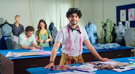 Indian young adult gen z man wear tape around neck learn new craft course look camera class room. Smart teen male boy pupil standing near desk table do casual pose staring cam enjoy day college place