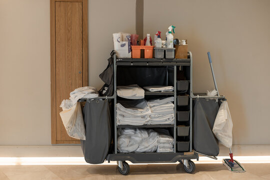 A cleaning cart filled with supplies, including towels, cleaning products, and a mop. The cart is parked against a wall in a well-lit area.