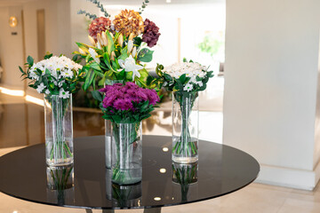 Three vases with colorful flowers on a black table in a bright indoor setting. The arrangement includes white, purple, and green flowers.