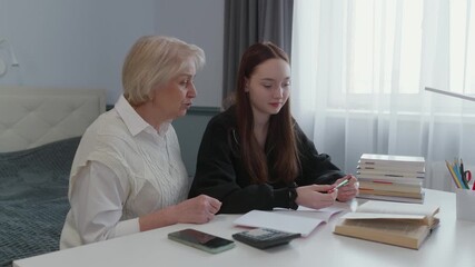 Experienced senior grandmother sitting at home with teenage granddaughter, offering educational support while helping with homework assignments and sharing generational wisdom