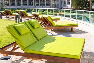 Lounge chairs with green cushions are arranged on a patio near a swimming pool. Surrounding palm trees and a modern hotel create a relaxing atmosphere.