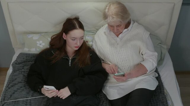 Teenage granddaughter guiding elderly grandmother through new mobile phone application, sharing digital knowledge while sitting closely together on home bedroom bed