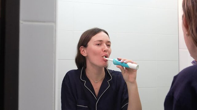 A woman's morning routine. A woman doing her morning hygiene routine. A woman brushing her teeth with an electric toothbrush in front of a mirror.