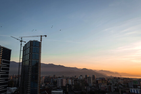Sunset view over Batumi with a high-rise under construction, cranes, mountains and sea in soft golden light. Urban skyline blends with natural landscape, creating a calm evening atmosphere.