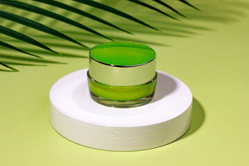 Jar of cosmetic cream on a white round shaped podium against light green background with tropical leaves