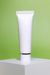 close up of empty white cream tube on a white square shaped podium against light green background. cosmetic mockup display