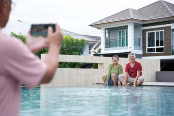 Senior couple posing by the pool while being photographed, Happy elderly couple relaxing by the poolside at home, Older couple smiling for a photo during a poolside moment