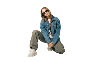 Young adult woman kneeling in studio wearing denim jacket and cargo pants, casual streetwear fashion with sunglasses and chunky sneakers for modern urban style lookbook