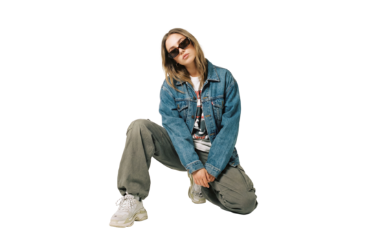 Young adult woman kneeling in studio wearing denim jacket and cargo pants, casual streetwear fashion with sunglasses and chunky sneakers for modern urban style lookbook
