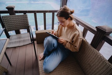 A young woman sits on a sunny veranda, drawing on a graphic tablet. Captures creative work process of a digital artist and designer in peaceful outdoor setting