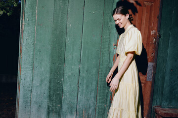 Fototapeta premium Casual portrait of a woman in a light dress walking beside a weathered doorway in an outdoor setting, with soft daylight and a vintage ambiance.