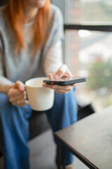 Person holding a smartphone and a coffee cup near a window in a bright cafe, with a soft depth of field. Concept modern communication, morning routine, comfort