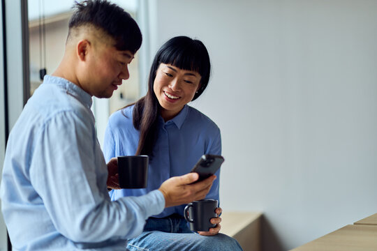 Asian Friends in Modern Workspace Sharing Coffee and Smartphone Conversation During Break At Office