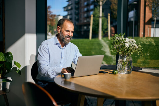 Businessman Working On Laptop At Cozy Café Table With Coffee, Flowers, And Sunlit View