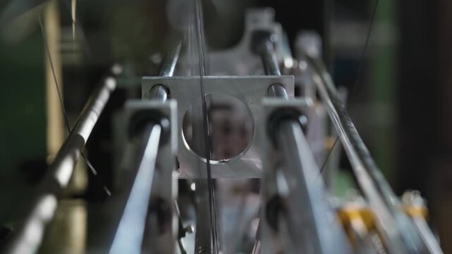 Camera moves forward through aligned metal rods of a weaving machine while textile threads pass between them, creating a shallow depth effect and dynamic industrial atmosphere