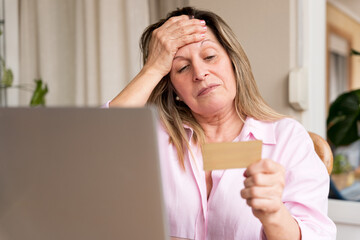 Stressed senior woman holding credit card, worrying about financial problems and online debt using laptop