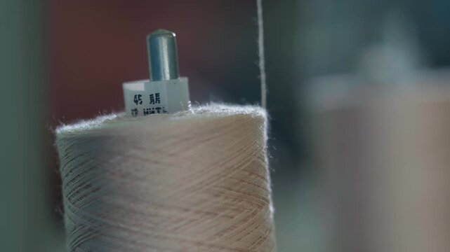 Soft close shot shows a beige thread spool with a single fiber rising toward the textile machine, highlighting delicate texture, gentle lighting and precise industrial setup