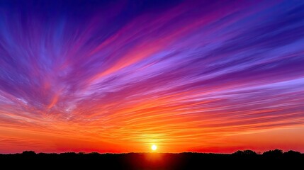 Obraz premium A vibrant sunset with streaks of orange, purple, and blue across the sky. The sun is setting behind a dark silhouette of trees.