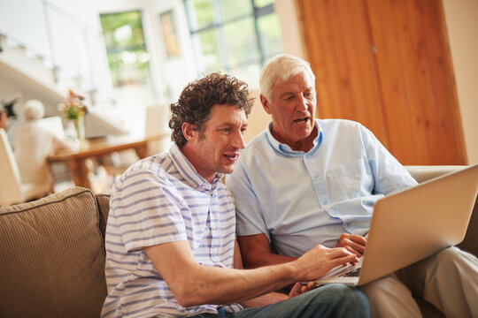Senior dad, man and help with laptop on sofa for web search, movie choice or bonding at family home. People, father and son with computer, typing and streaming with guide on couch at apartment
