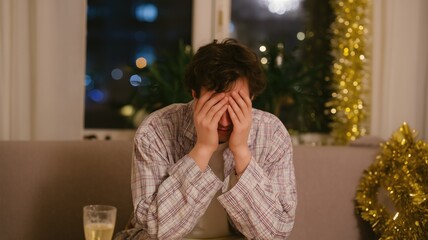 A young man sitting on the couch at home, covering his face with hands and crying, because he is alone for a New Year's eve party
