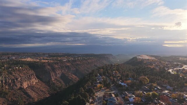 Wide aerial shot featuring residential town development on a forested mesa top. The Los Alamos airstrip is visible in the background, set against the steep, dramatic canyon gorge at sunrise.