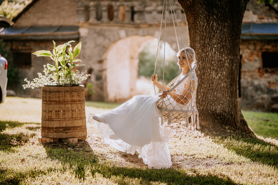 Bride in white dress swinging gently on a rustic swing beneath a tree, surrounded by greenery and a wooden barrel, capturing a serene outdoor wedding atmosphere