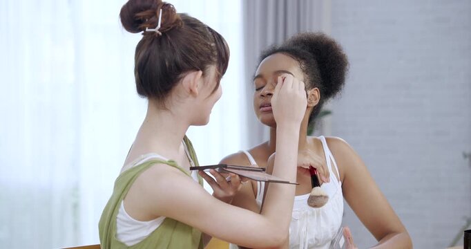 asian teenage beauty vlogger using makeup brush applying eyeshadow on african american teenage girl eyelid while smiling during home tutorial shoot as part of cosmetic influencer routine lifestyle