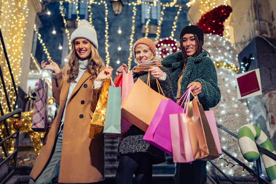 Happy young women Christmas shopping with festive lights, colorful gift bags, joyful holiday spirit and winter fashion
