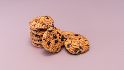 A mix of stacked and scattered delicious, golden-brown chocolate chip cookies is centrally placed on a clean, light mauve background, suggesting an informal, inviting snack.