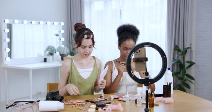 asian teenage girl holding mirror smiling while african american teenage girl holding brush during slowmotion skincare tutorial shoot at home setup showing joyful cosmetic influencer lifestyle routine