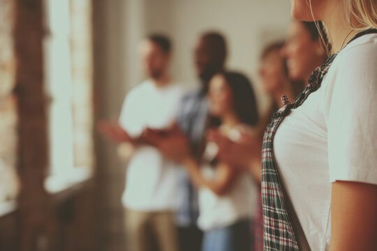 Young woman in foreground with group of diverse people clapping in blurred background, warm indoor light and supportive atmosphere