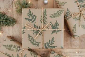 Rustic holiday gifts wrapped in kraft paper with green fern pattern and twine on wooden table. Eco-friendly Christmas present styling, warm lights.