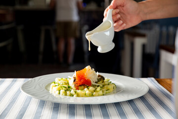 Chef pouring creamy sauce onto an elegant gourmet seafood appetizer with red caviar and chopped vegetables on a white plate.