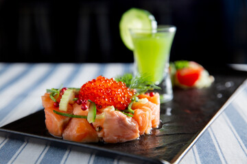 Gourmet salmon tartare with vibrant red caviar on a black plate. Served with fresh cucumber, dill, and a green shooter cocktail.