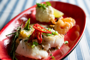 Gourmet plated white fish fillet with shrimp, cauliflower puree, chili, and fresh herbs on a red plate. Elegant seafood.